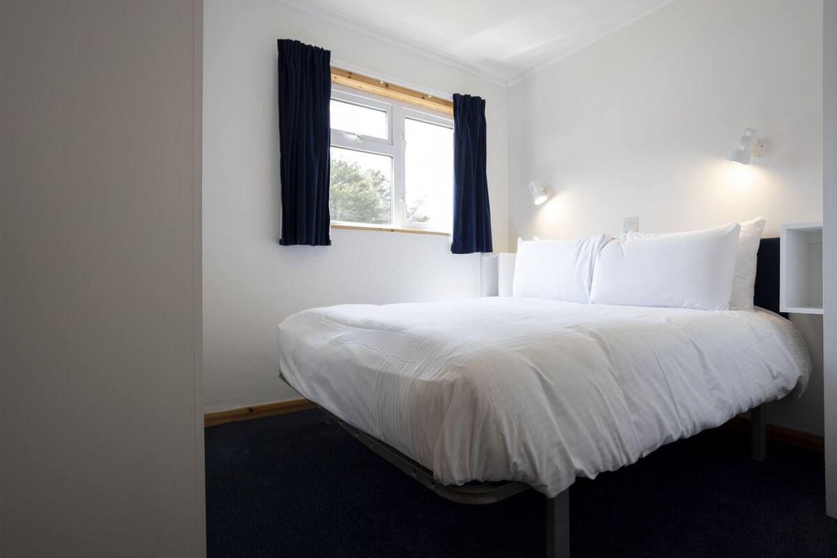 A neatly made bed with white bedding in a room with white walls, a window with dark curtains, and a bedside table with a lamp.