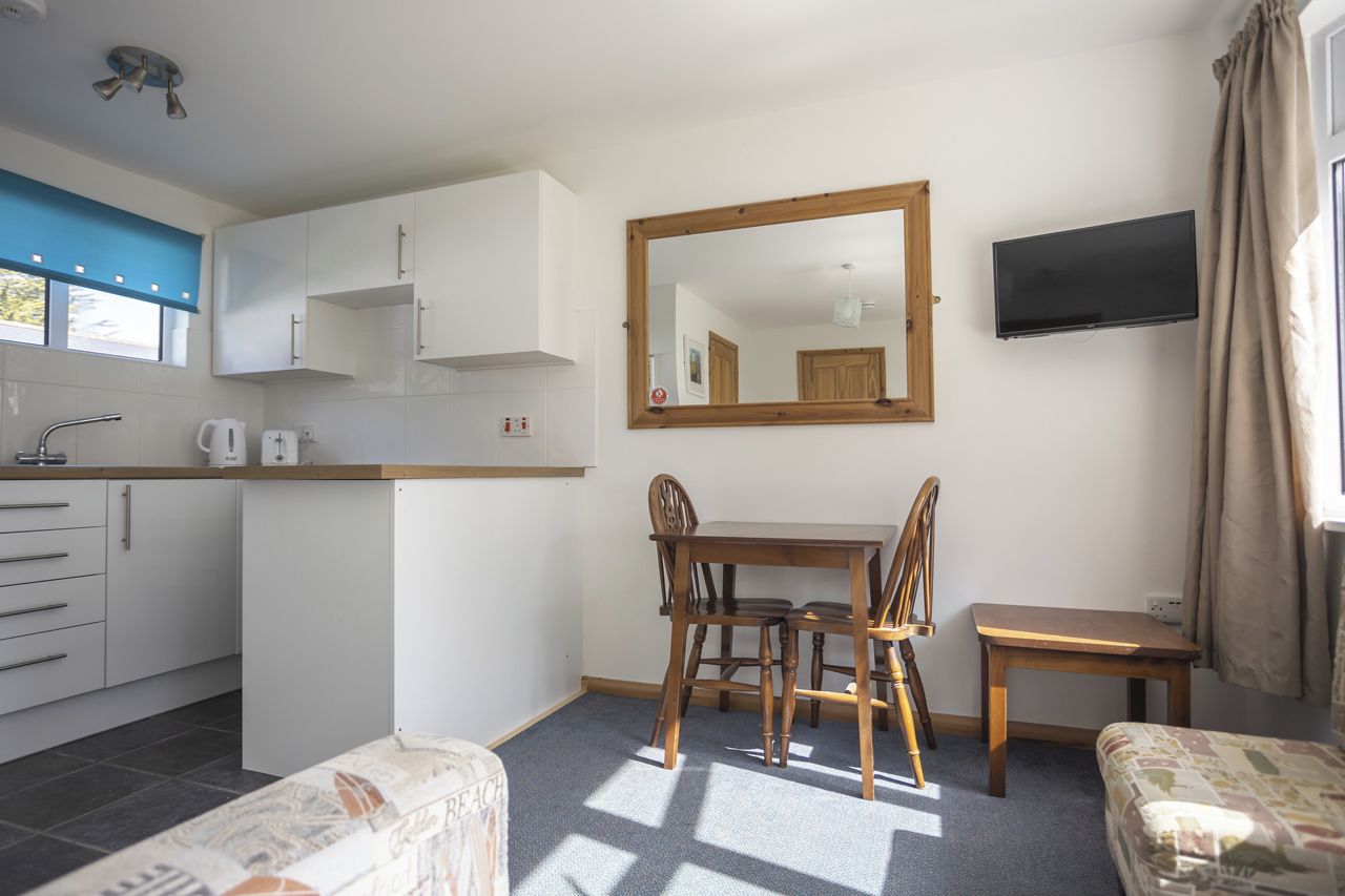 A compact kitchen with white cabinets, a wooden dining table, and a flat-screen TV mounted on the wall.