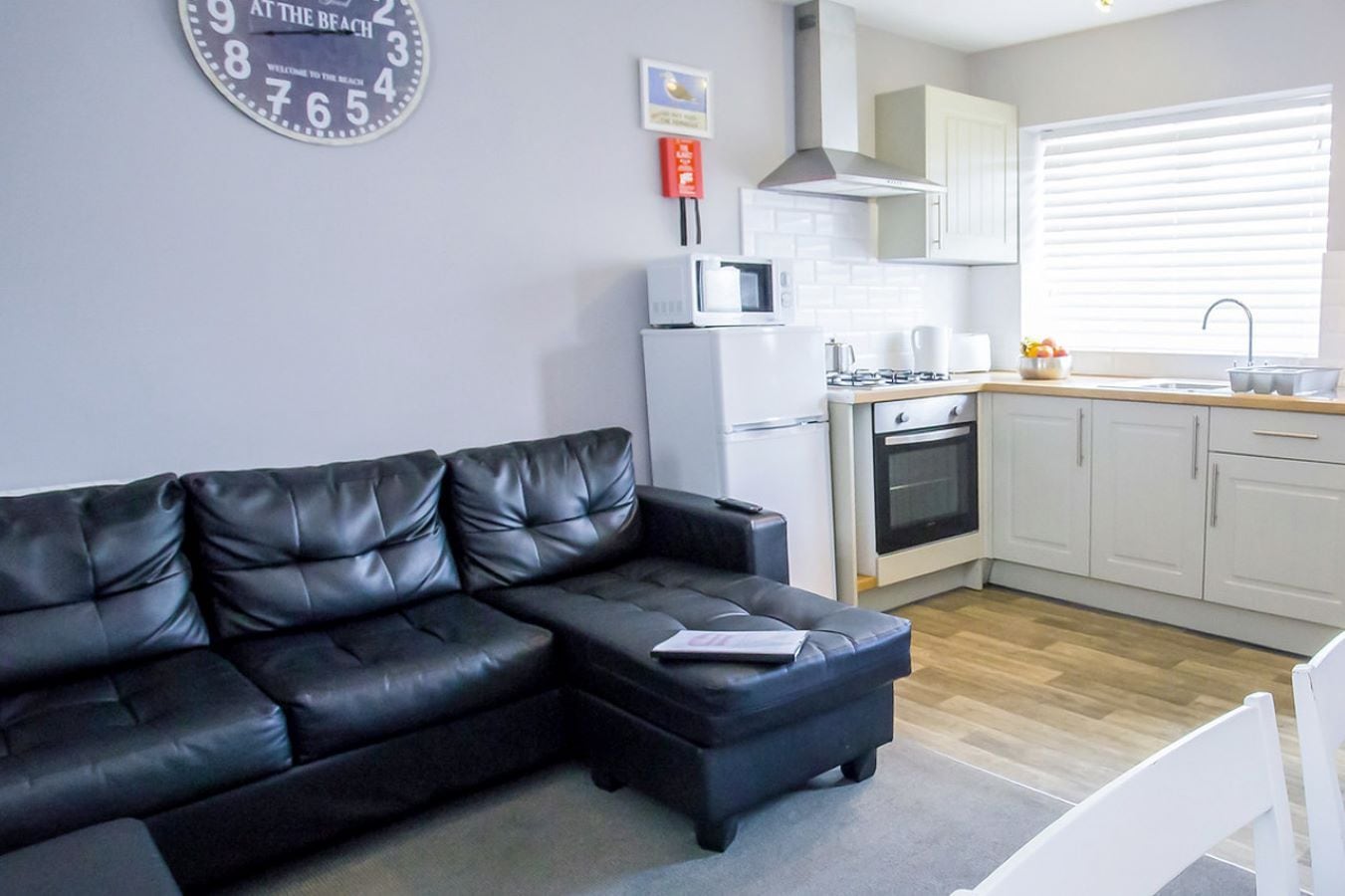 A modern living room with a black leather sofa, a white kitchen area including a microwave, oven, and cabinets, and a clock on the wall.