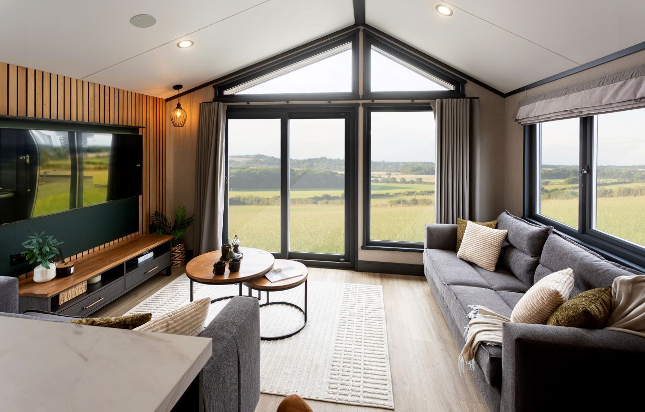 A modern living room with a large TV, a sofa, a coffee table, and a view of the countryside through large windows.