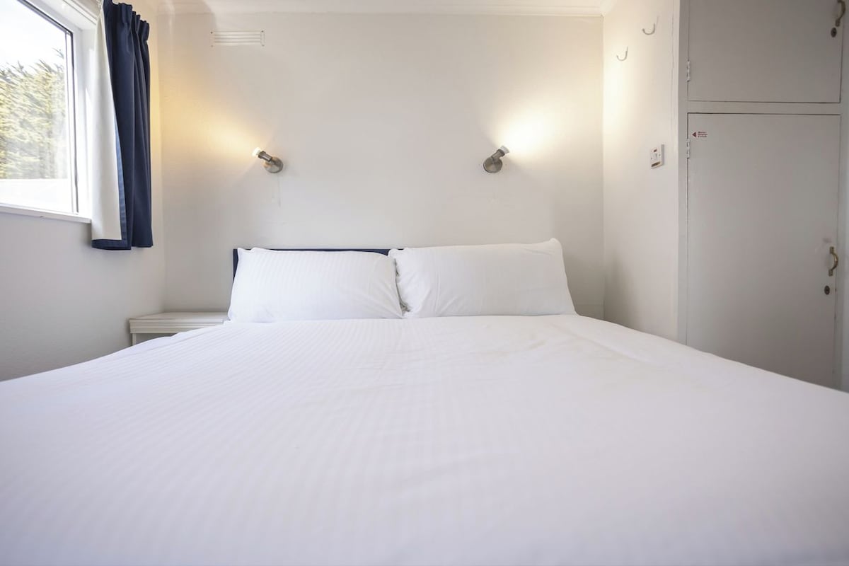 A large bed with white linens, two wall-mounted light fixtures, a window with curtains, and a door.