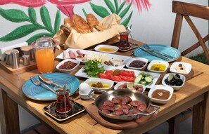 Uma mesa de pequeno-almoço com diversos alimentos, incluindo pão, queijo, tomates, azeitonas e uma travessa de carne.