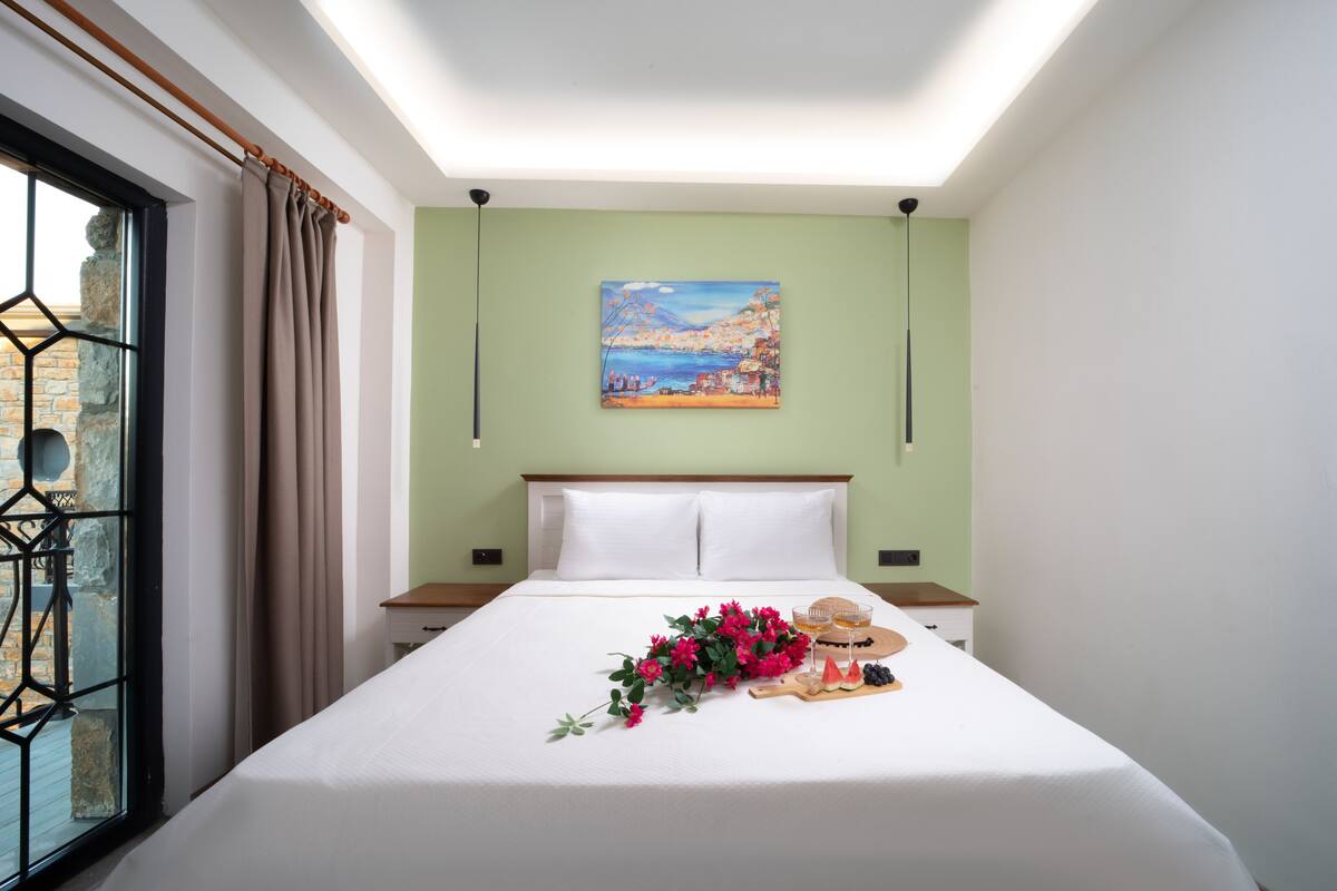 A neatly made bed with a white sheet, a painting on the wall, and a window with curtains.