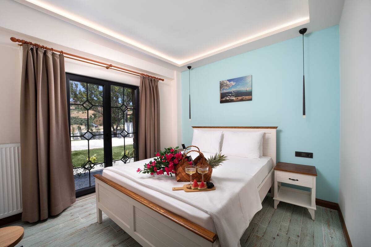 A bedroom with a bed, a nightstand, a window with curtains, and a view of the outdoors.