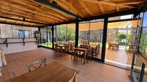 Un amplio espacio de comedor al aire libre con mesas y sillas de madera, grandes ventanales y vistas a un jardín.