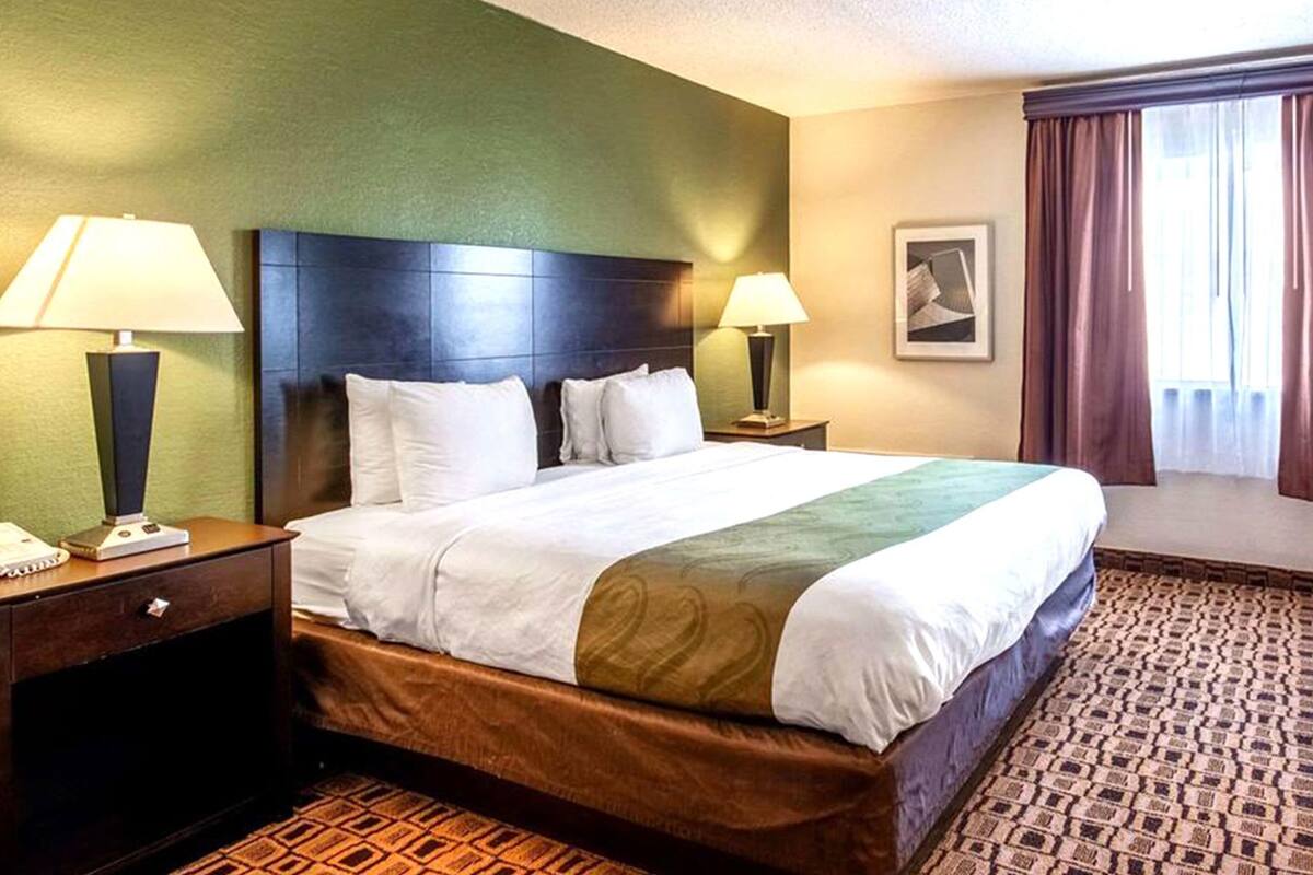 Habitación de hotel con una cama grande, dos mesitas de noche, una lámpara, un cuadro enmarcado y una ventana con cortinas.