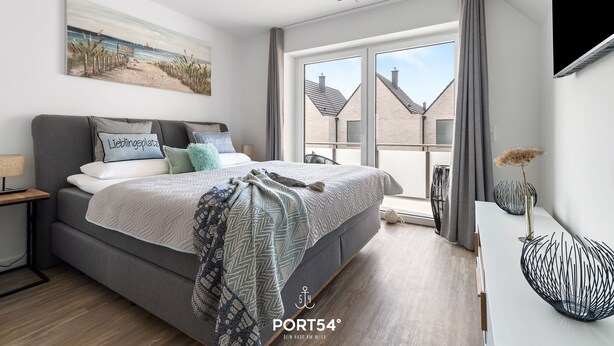Une chambre moderne avec un grand lit, une table de chevet, un tableau représentant une plage et une vue sur des immeubles par une grande fenêtre.