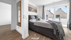 Une chambre à coucher moderne avec un grand lit, un tableau au mur et une vue sur les immeubles par la fenêtre.