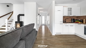 Un salon moderne avec un canapé gris, un poêle à bois et une cuisine dotée de meubles blancs et d’un plan de travail en bois.