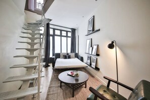 A compact room with a bed, a round coffee table, a chair, a staircase, and a window with curtains. - Triplex-6Pers-Porte Maillot-ArcdeTriomphe-Paris17 (Paris)