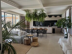 Un salon moderne comprenant un canapé, une table basse et des fauteuils, un coin cuisine avec une table à manger, et de grandes fenêtres laissant entrer la lumière naturelle.