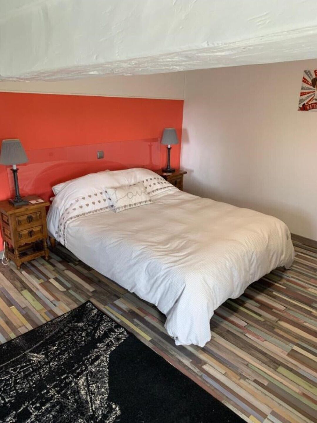 A single-bed room with a white bedspread, two bedside tables with lamps, a patterned rug, and a multicolored wooden floor.