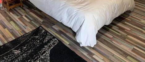A single-bed room with a white bedspread, two bedside tables with lamps, a patterned rug, and a multicolored wooden floor.
