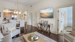 A spacious living area with a dining table, white chairs, a flat-screen TV, and a kitchen in the background.