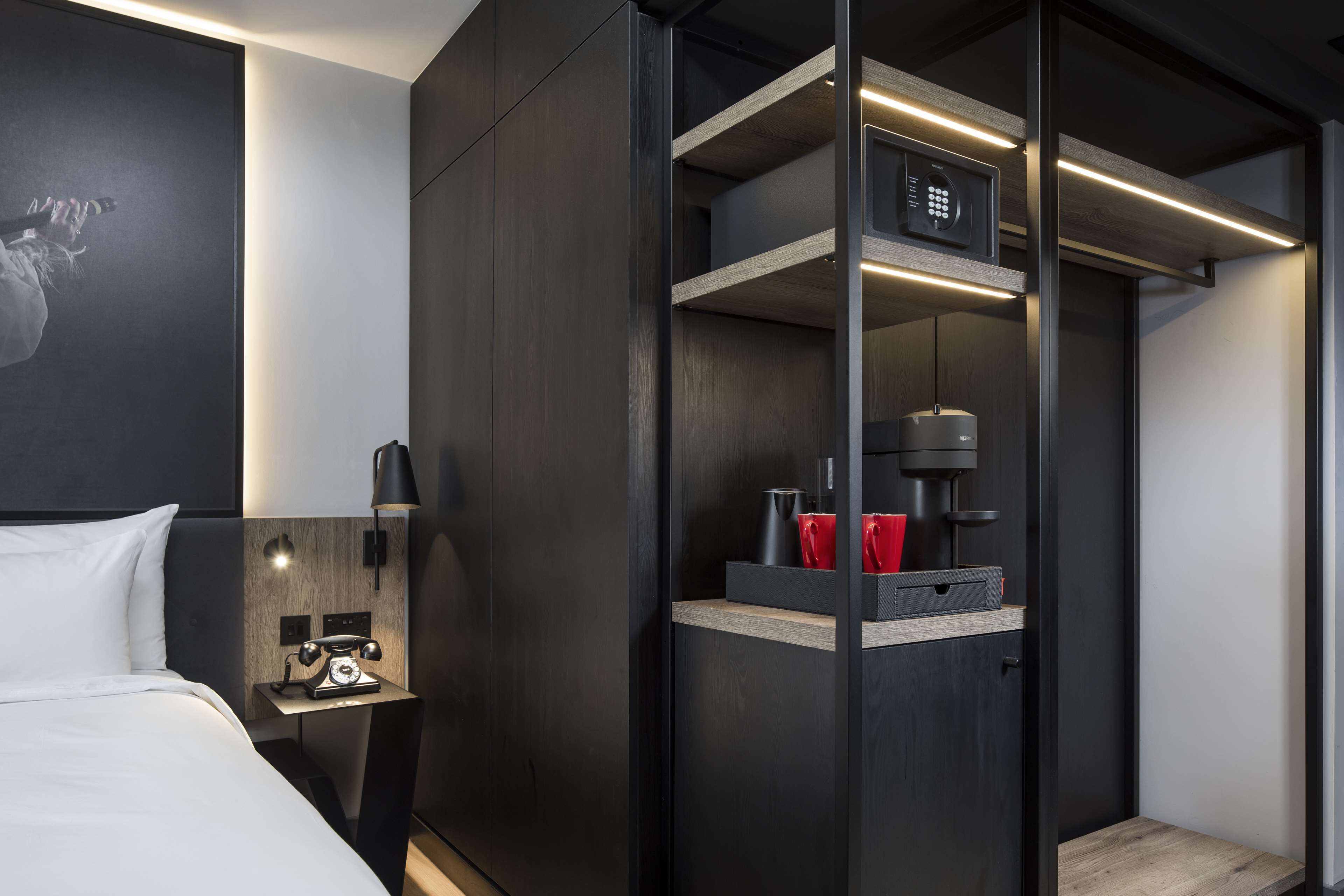 Una habitación de hotel moderna con cama, mesita de noche con teléfono antiguo, armario empotrado con cafetera y una pequeña estantería con vasos rojos.