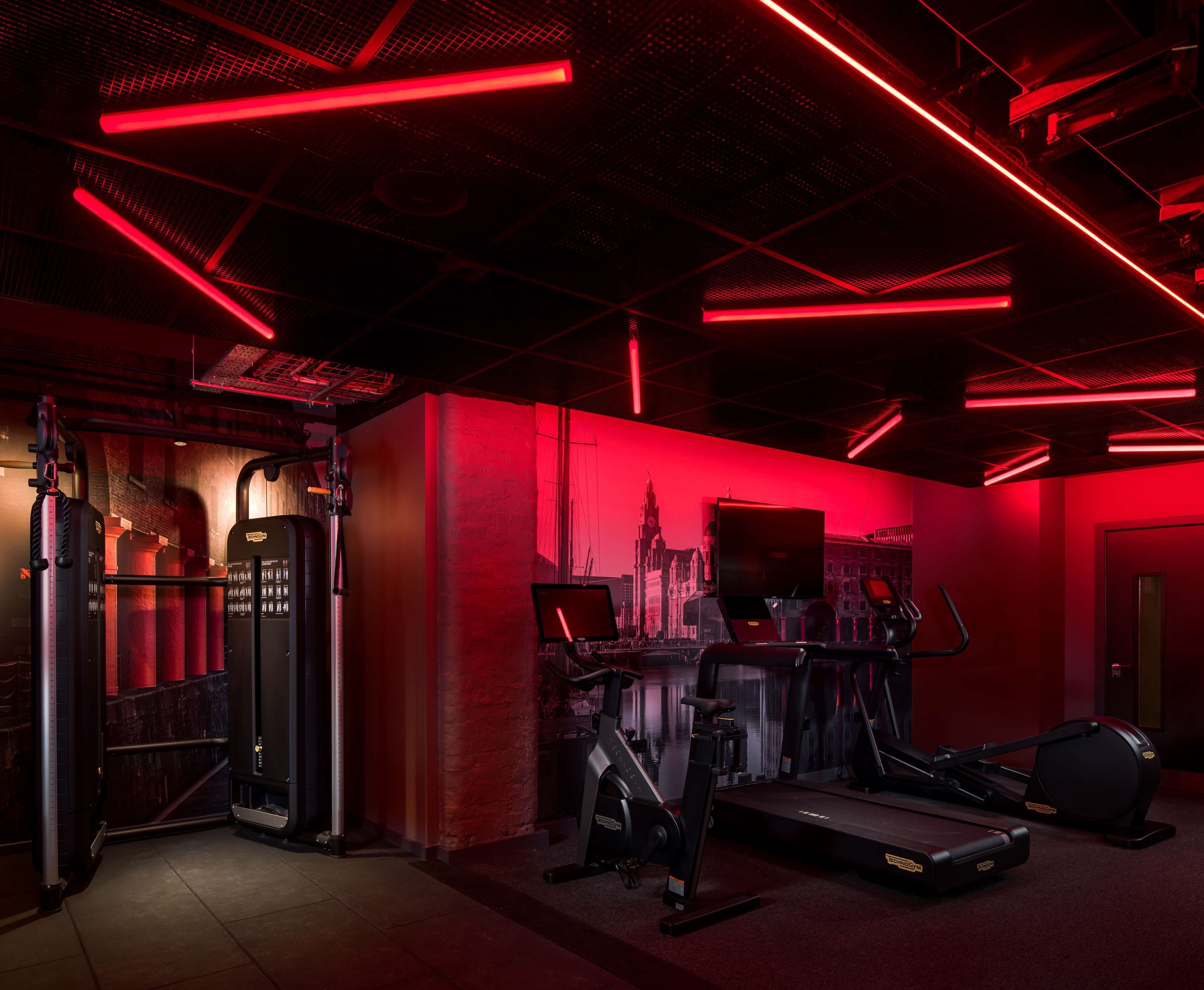 A gym with red lighting, exercise equipment, and a cityscape mural on the wall.