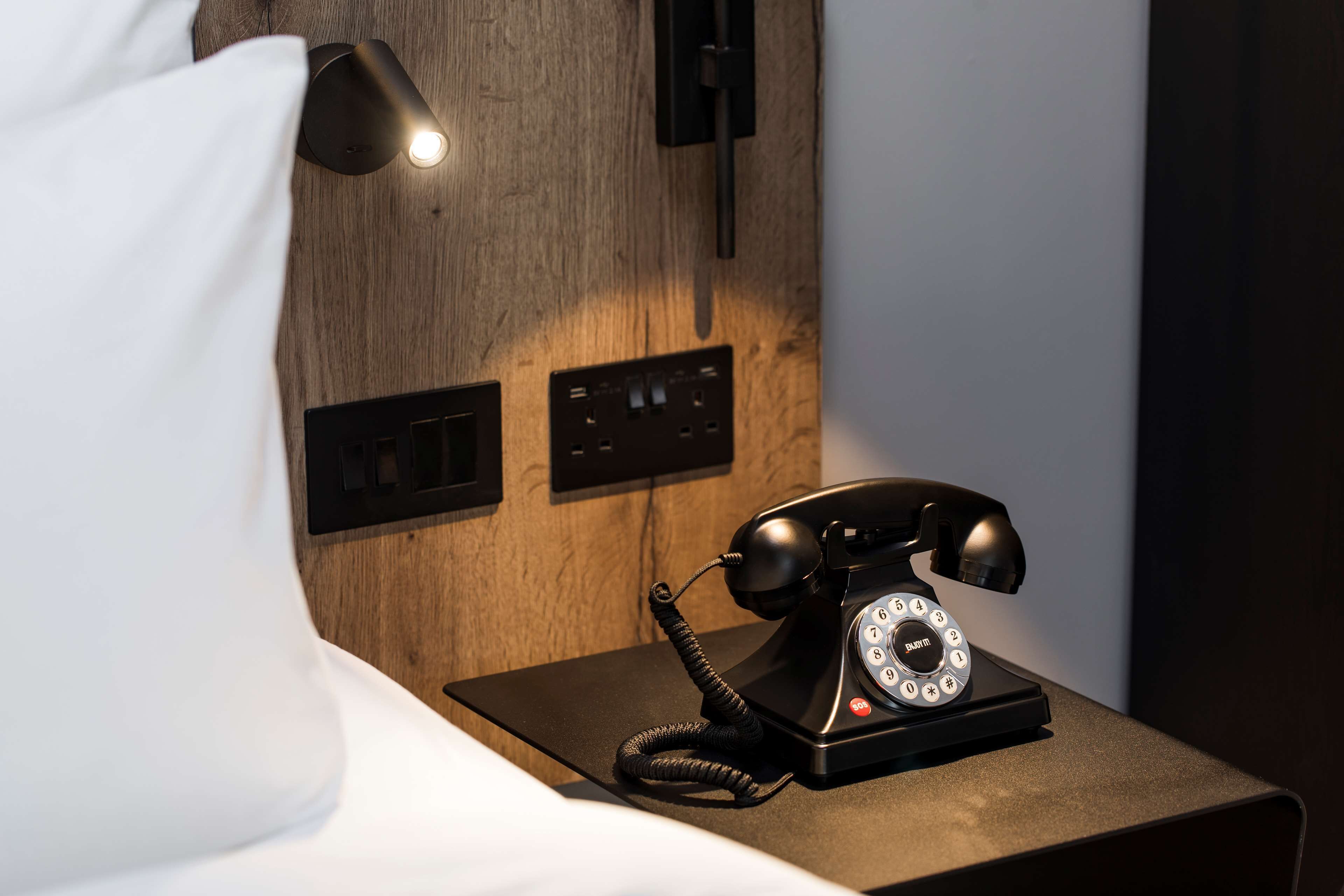 A wooden bedside table with a black rotary phone and a wall-mounted light fixture.