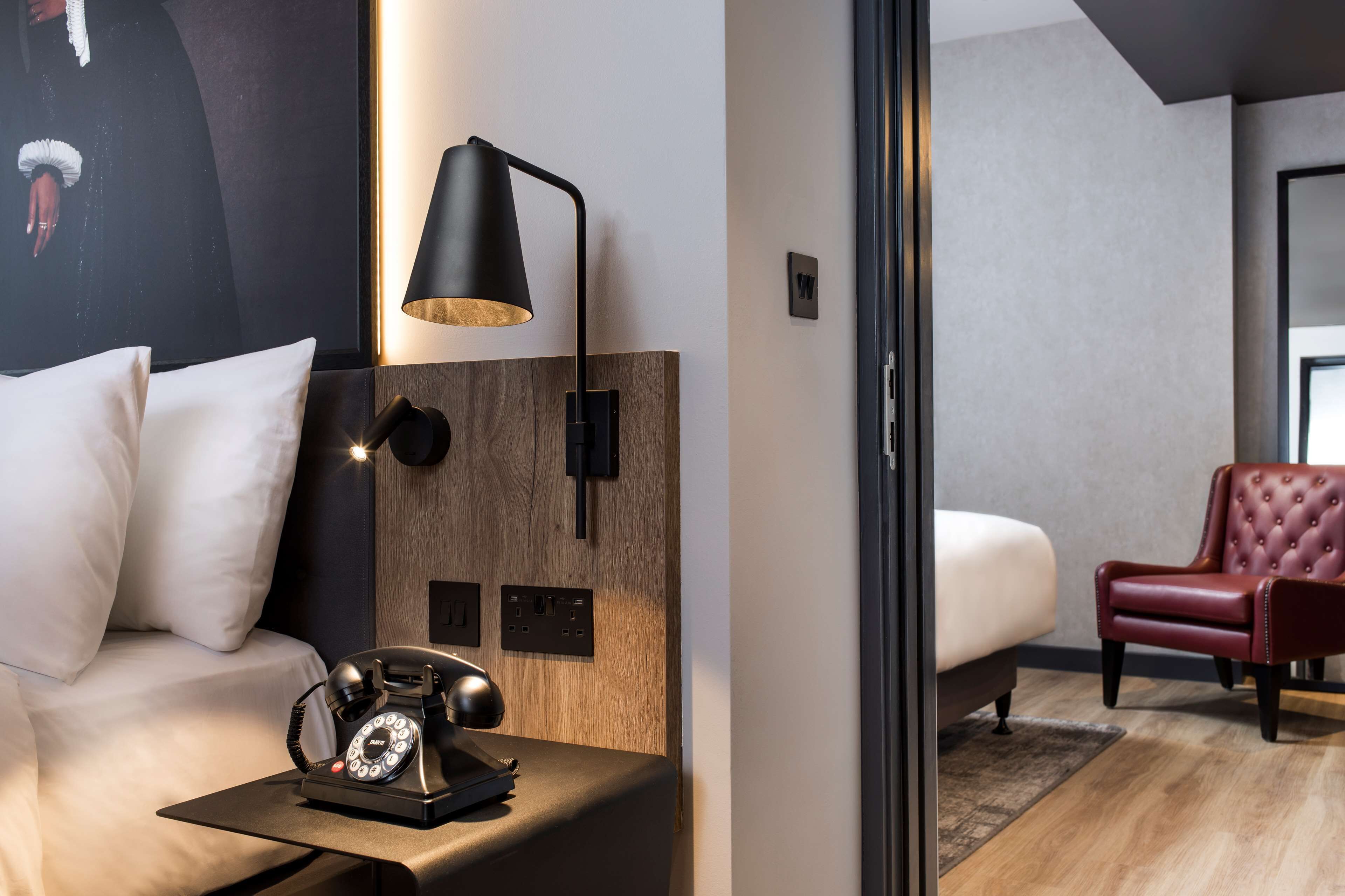A modern hotel room with a wooden headboard, a black telephone, a black desk lamp, a red leather armchair, and a white bed.