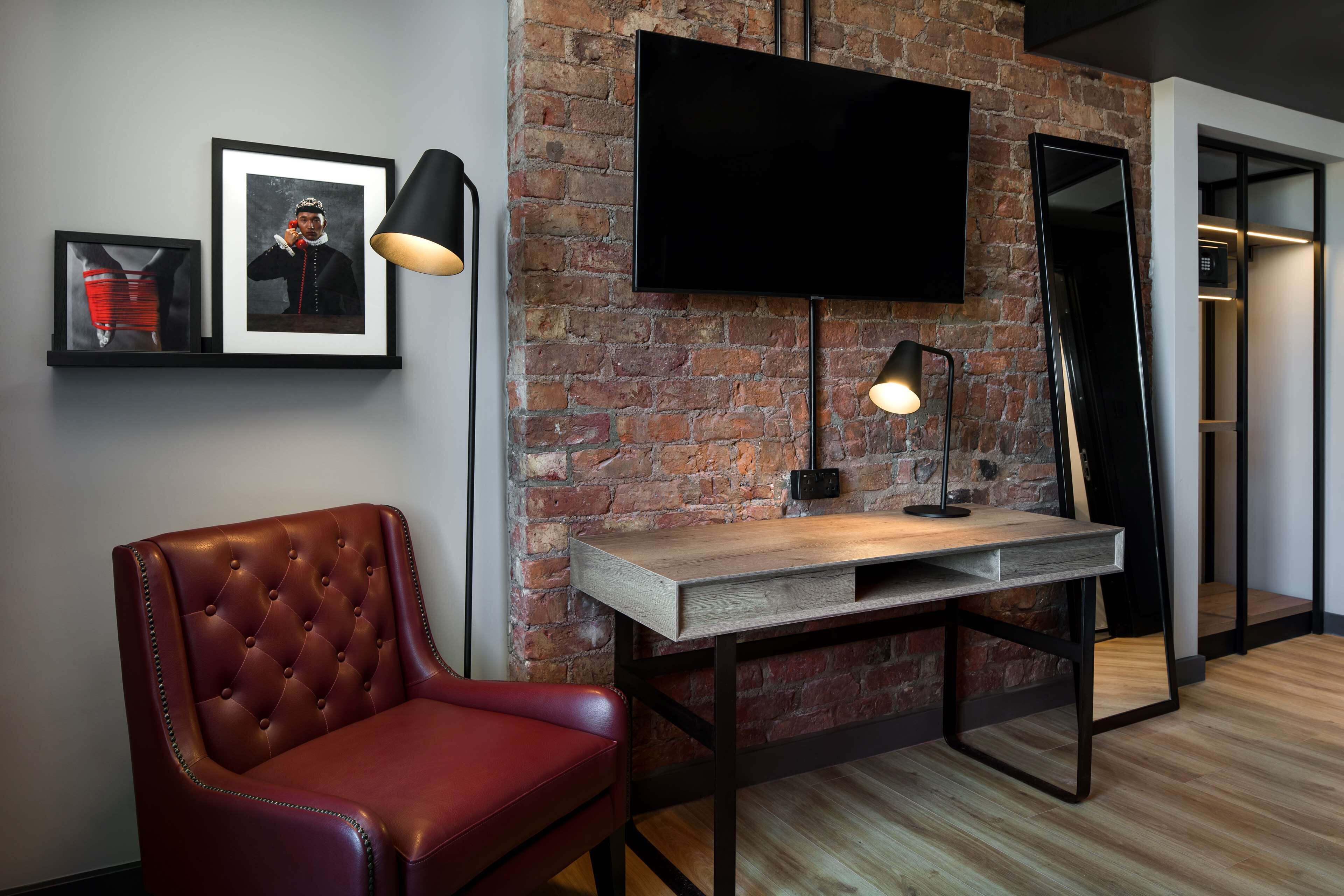 A modern office space with a brick wall, a flat-screen TV, a leather chair, a wooden desk, and a standing lamp.