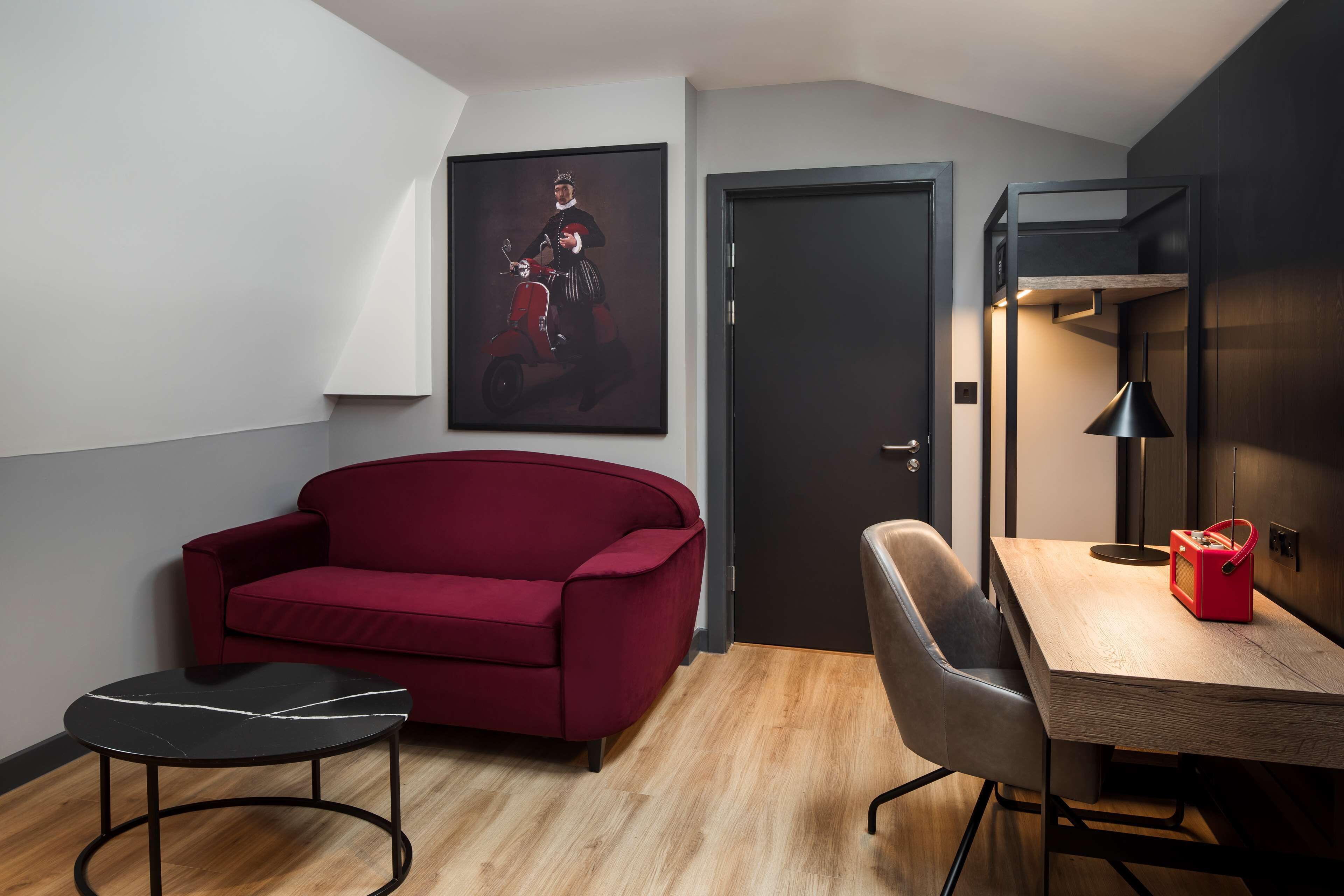 A modern room with a red sofa, a small round table, a wooden desk with a lamp, and a door.