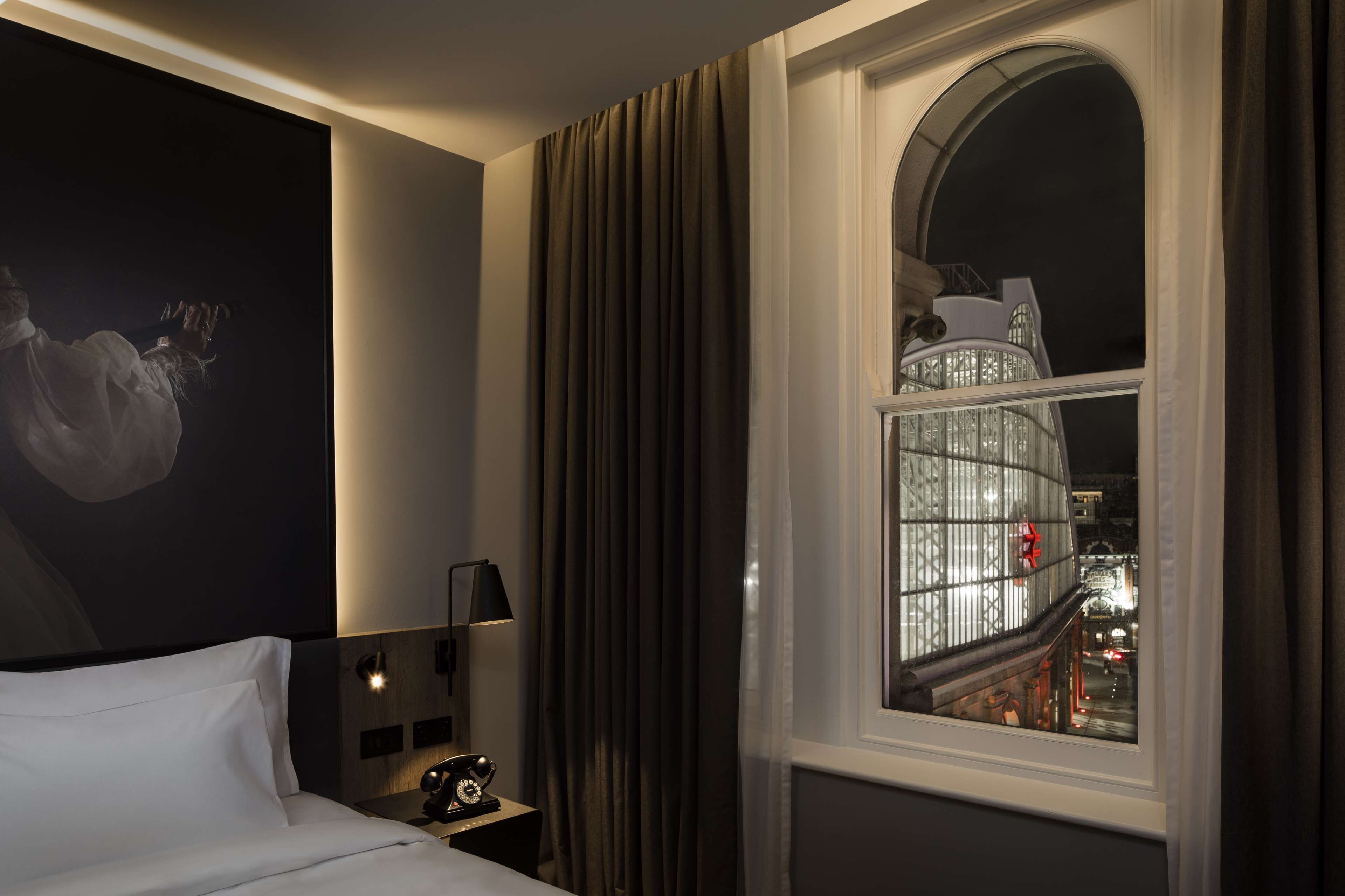 A hotel room with a bed, a nightstand, a lamp, a large window with curtains, and a cityscape view at night.