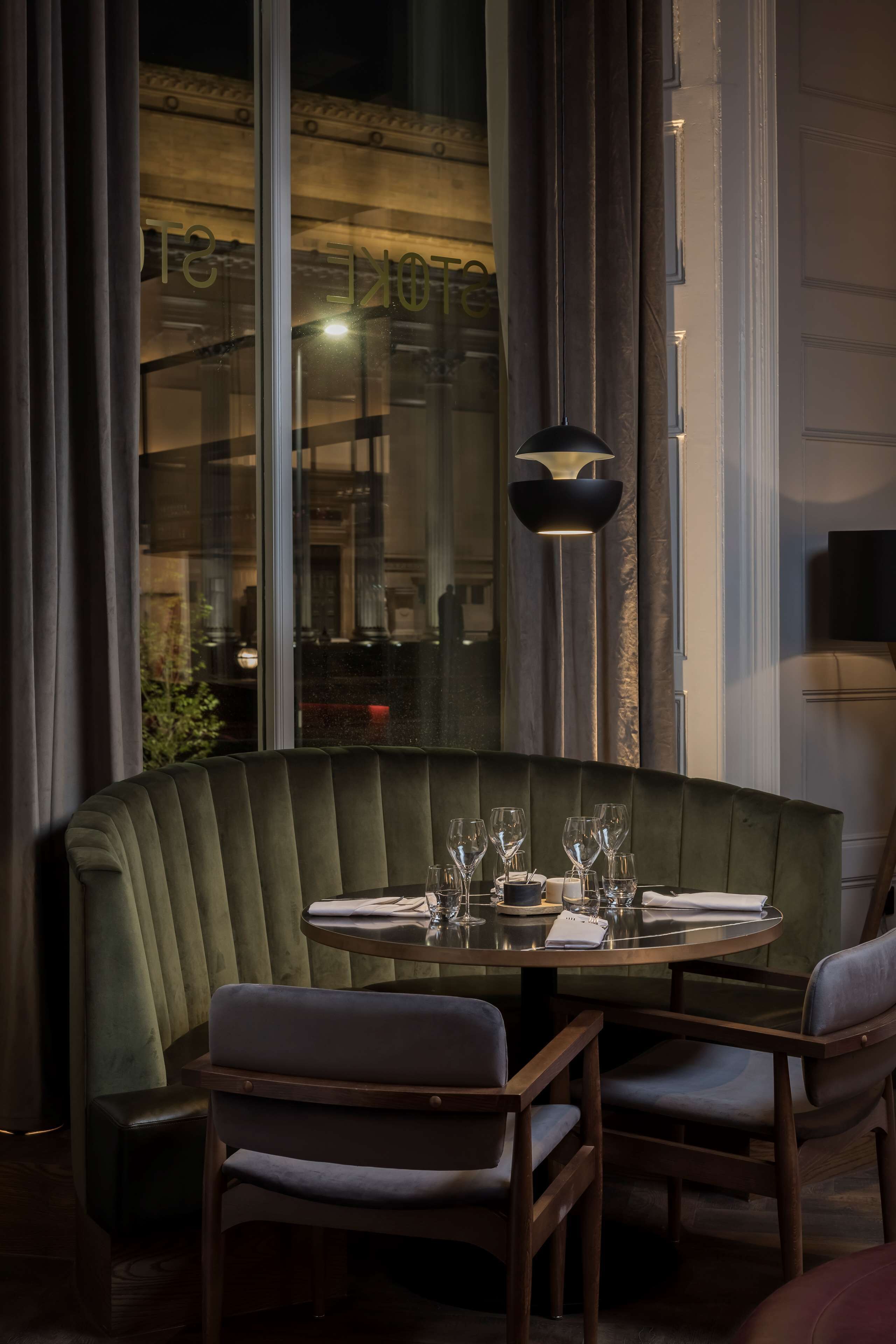 A corner dining area with a green upholstered booth, wooden chairs, a round table set with glasses and plates, and a pendant light.