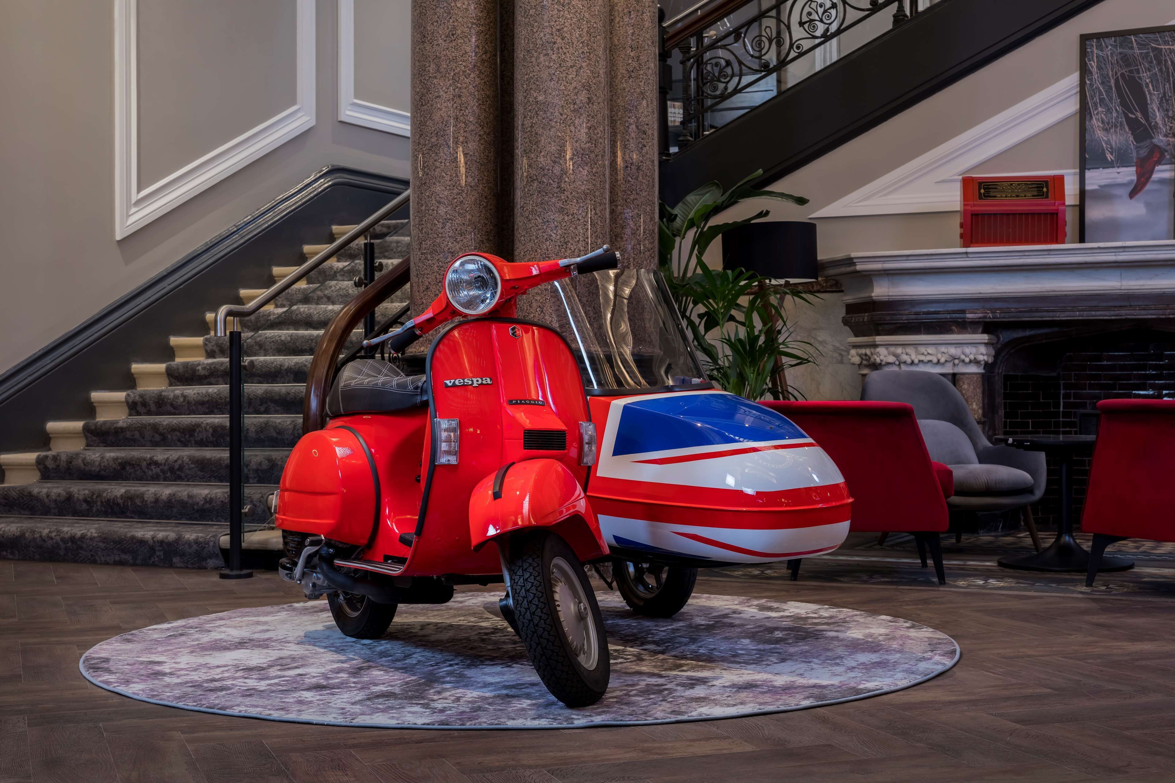 A red Vespa scooter with a Union Jack design parked on a patterned rug in a lobby area with a staircase and modern furniture.
