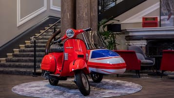 A red Vespa scooter with a Union Jack design parked on a patterned rug in a lobby area with a staircase and modern furniture.