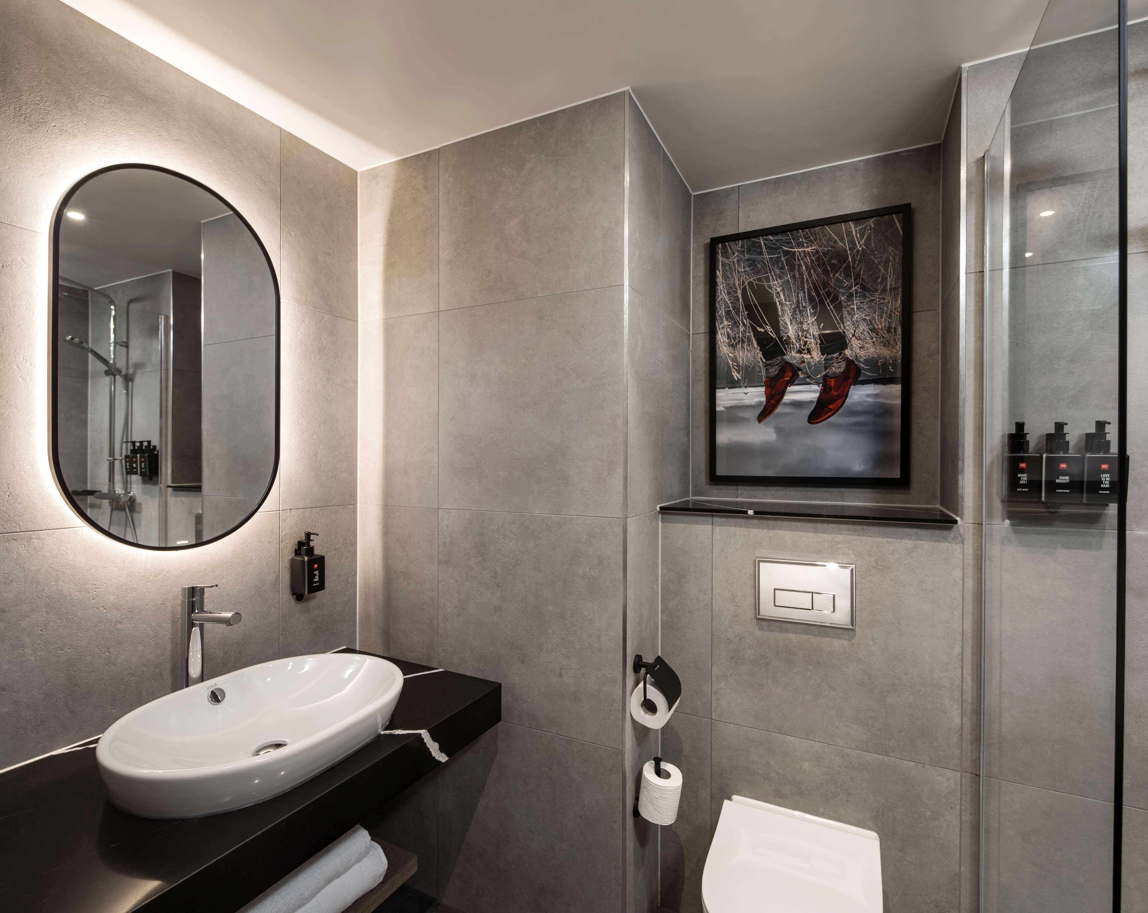 A modern bathroom with a large oval mirror, a white sink, a toilet, and a shower area.