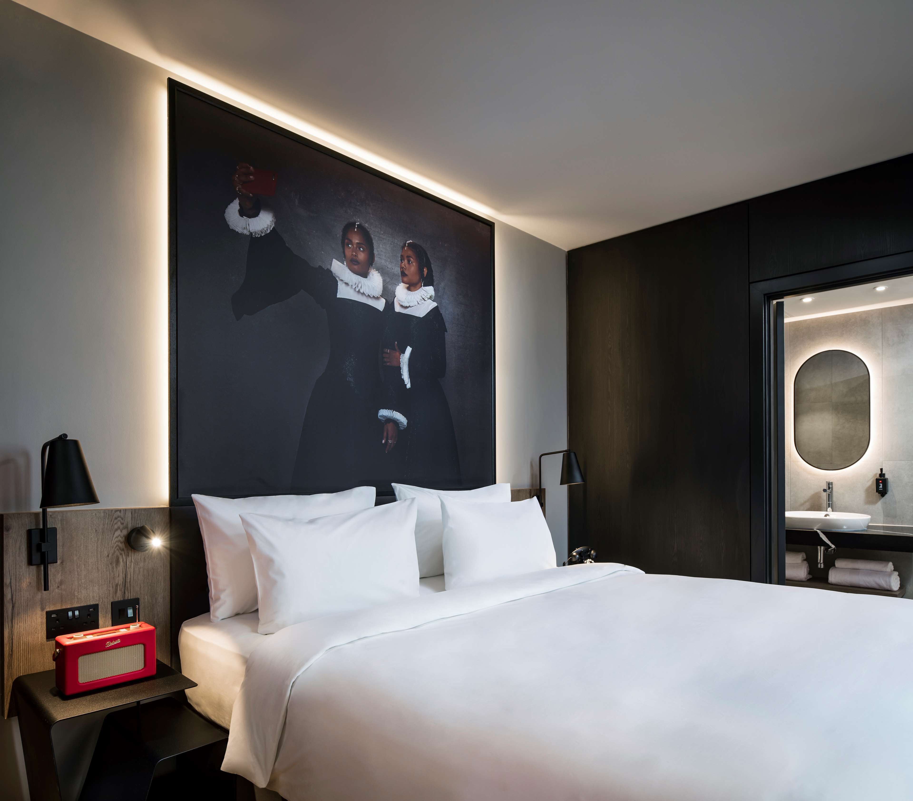 A modern hotel room with a large bed, a nightstand with a red radio, a wall-mounted lamp, and a large mirror with a round frame.