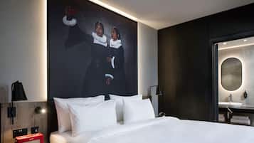 A modern hotel room with a large bed, a nightstand with a red radio, a wall-mounted lamp, and a large mirror with a round frame.