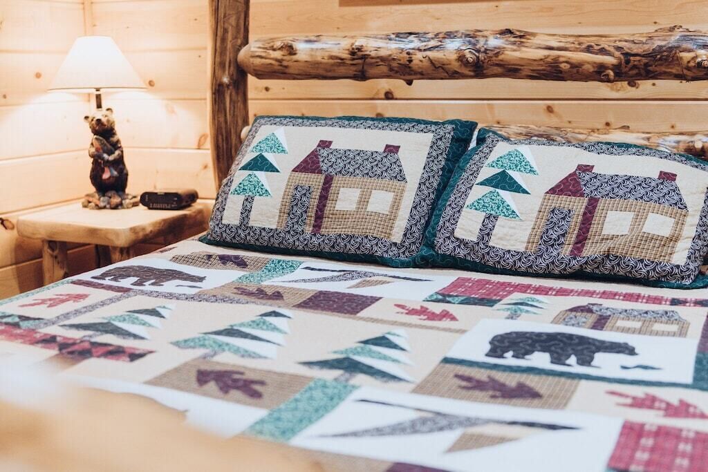Ein Holzblockhaus-Interieur mit einer gesteppten Bettdecke mit Tiermotiven und Hausmustern, einer Lampe und einer Bärenfigur auf einem Nachttisch.