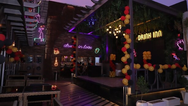 A modern bar area with neon signs, including "DRINK IN LOVE" and "DRU in love," and a stage with a backdrop of balloons. - DD Suites (Agra)