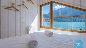 A bedroom with a bed, a window with a view of a lake and mountains, wooden wall panels, and a towel on the bed.