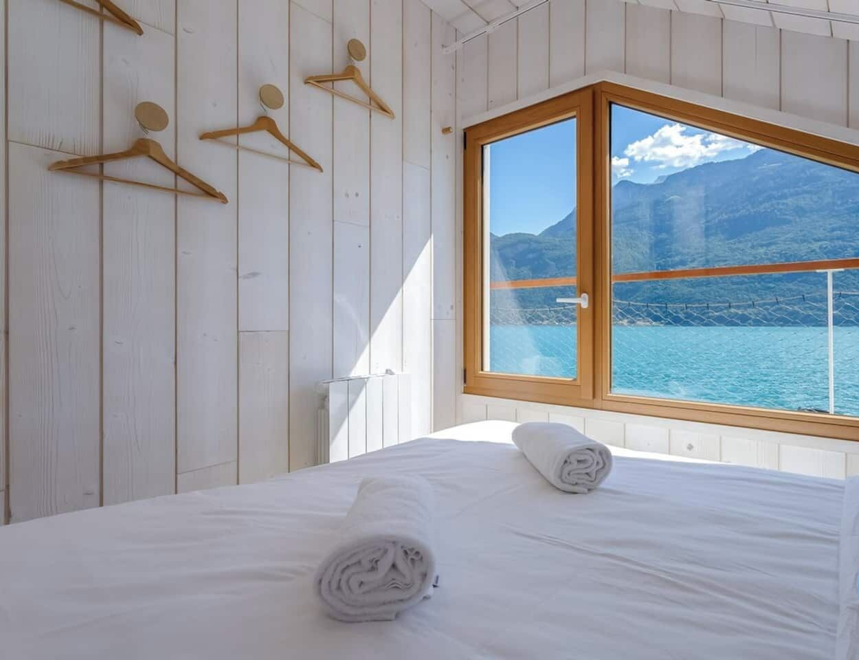 Een slaapkamer met een bed, een raam met uitzicht op een meer en bergen, houten wandpanelen en een handdoek op het bed.