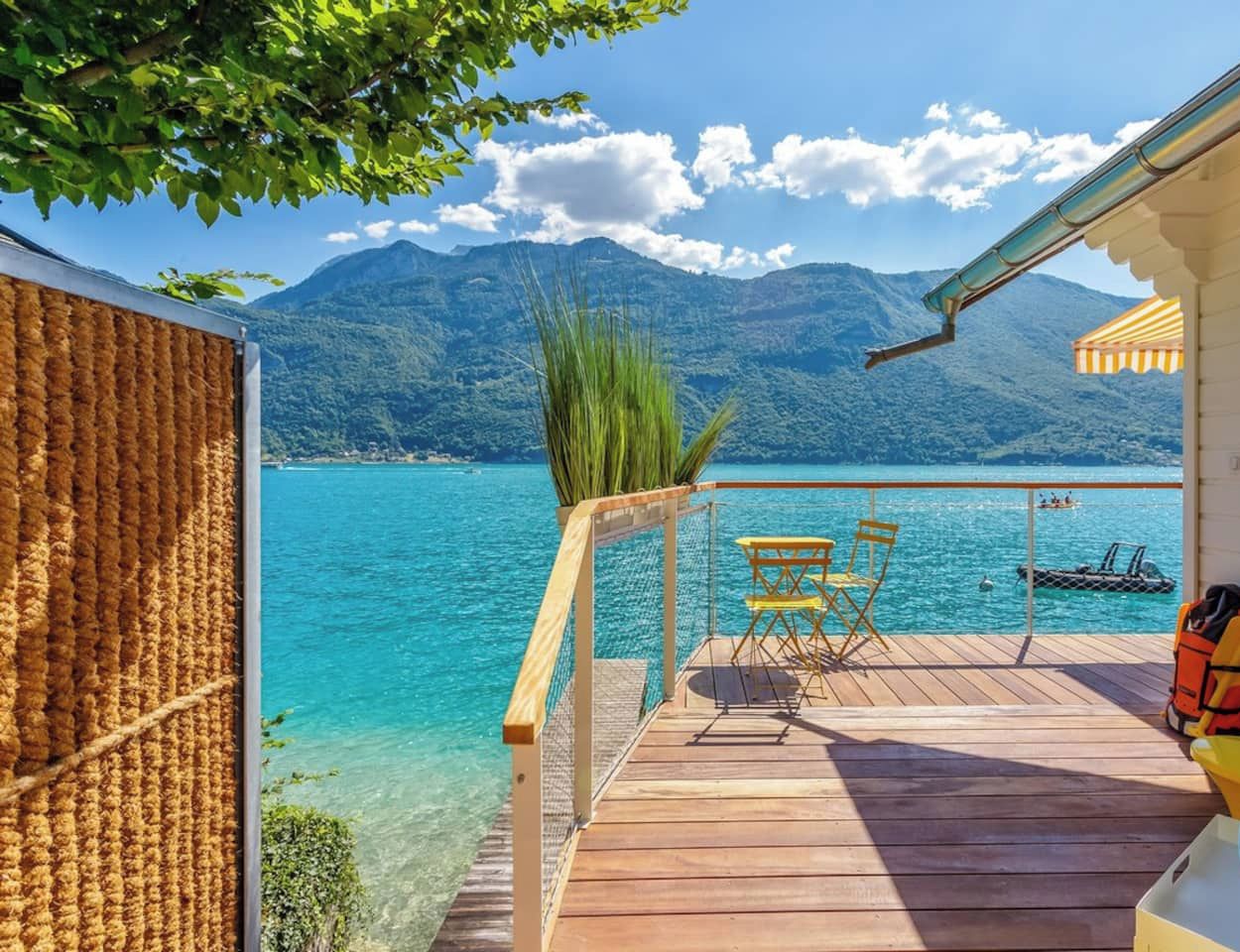 A wooden deck with a view of a lake and mountains, a table and chairs, and a life jacket on the right.