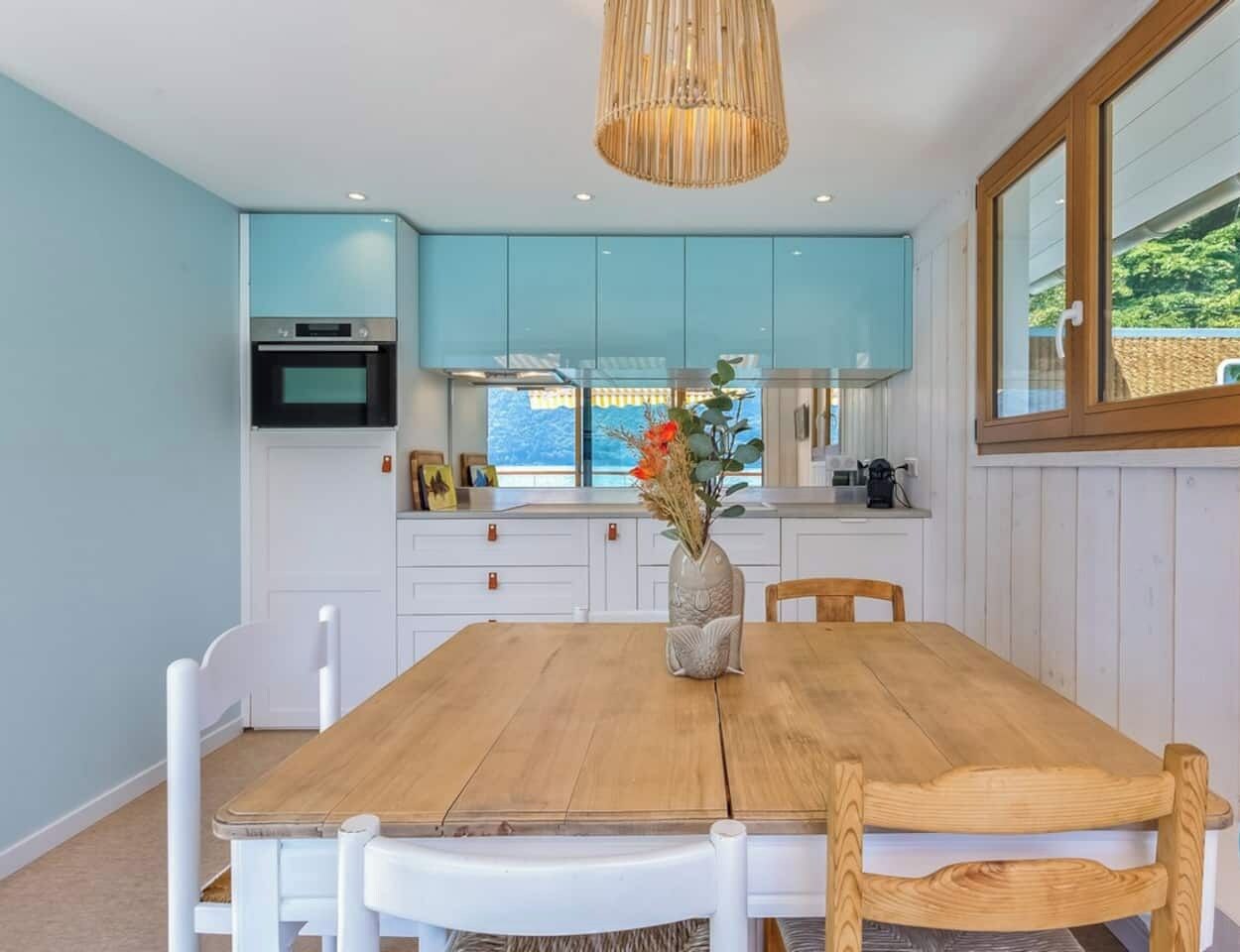 Een eetgedeelte met een houten tafel en stoelen, een vaas met bloemen en lichtblauwe kasten.
