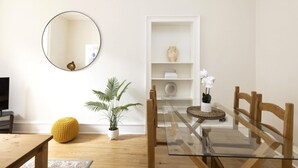 Uma sala de jantar com mesa de vidro, cadeiras de madeira, um banquinho amarelo, uma planta em vaso, uma prateleira branca com objetos decorativos e um espelho redondo na parede.