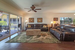 A spacious living room with a large sectional sofa, a coffee table, and a ceiling fan. There are two framed pictures on the wall, and a sliding glass door leads to an outdoor patio area.