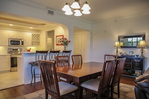 Uma sala de jantar com mesa e cadeiras de madeira, uma cozinha com eletrodomésticos modernos e uma sala de estar com luminária e objetos decorativos.
