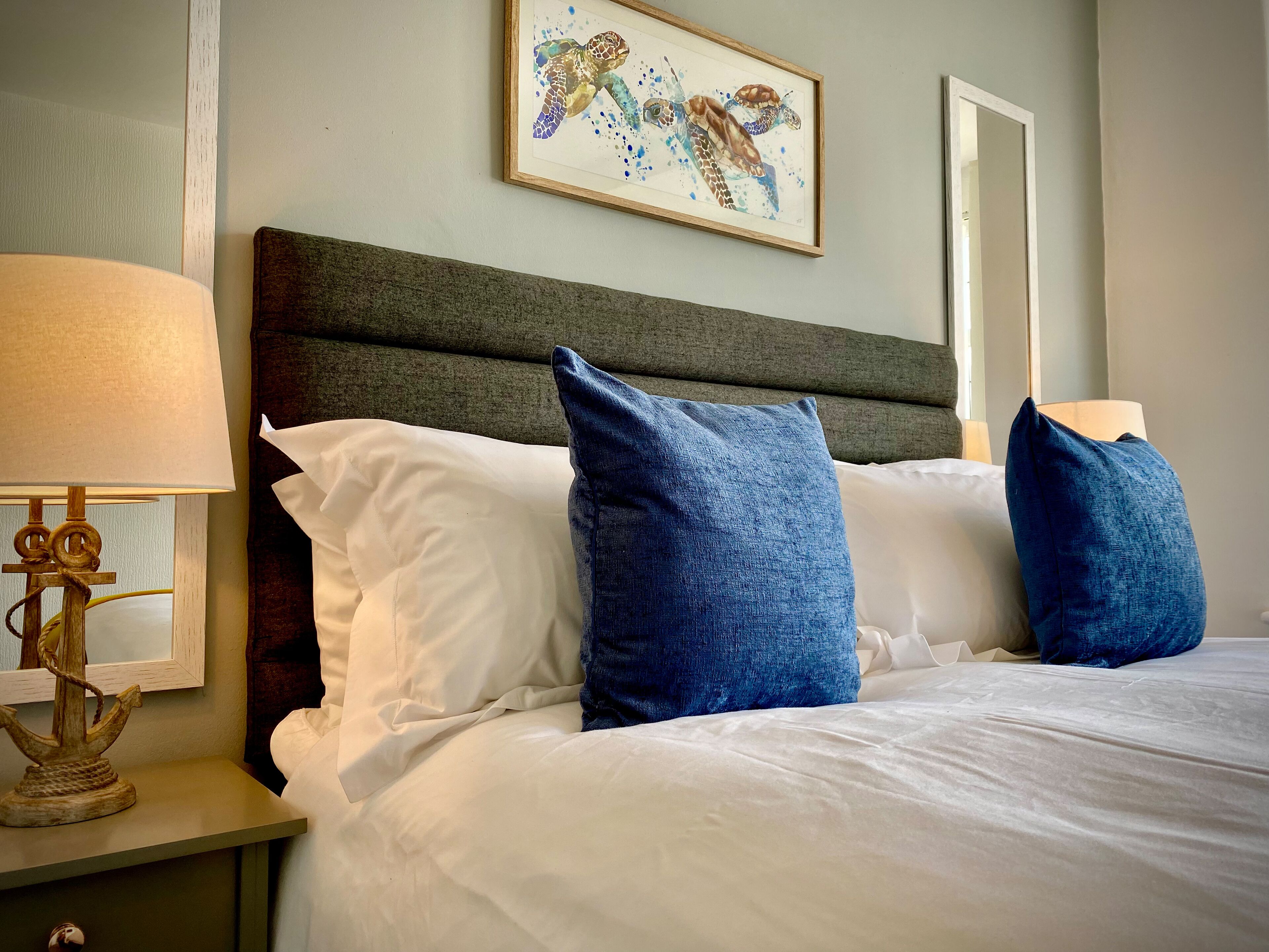 A neatly made bed with white and blue pillows, a wooden nightstand with a lamp, and a framed artwork of sea turtles on the wall.