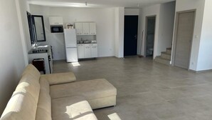Un vaste espace de vie avec un canapé d’angle beige, une kitchenette dotée de meubles blancs et une porte sombre en arrière-plan.