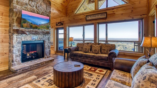 Un intérieur de chalet chaleureux avec une cheminée en pierre, de grandes fenêtres offrant une vue imprenable et un coin salon confortable.