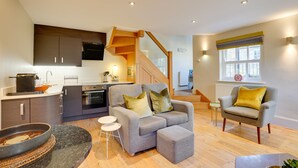 A modern living room with a grey sofa, a small armchair, and a dining area with a kitchen in the background. - Garden Stable: Cosy Stone Cottage with Parking (Alnmouth)