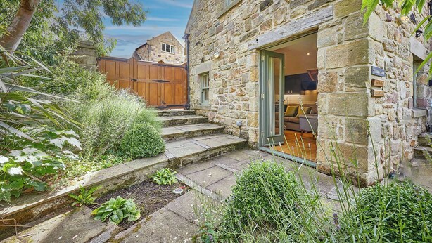 A stone building with a wooden door, a staircase leading to a porch, and a garden area with various plants. - Garden Stable: Cosy Stone Cottage with Parking (Alnmouth)
