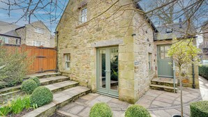 A stone house with a green door, a wooden fence, and a paved garden area with trimmed bushes and a small tree. - Garden Stable: Cosy Stone Cottage with Parking (Alnmouth)