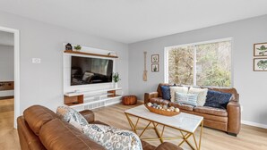 Ein Wohnzimmer mit braunem Ledersofa, einem weißen Couchtisch und einer braunen Lederliege. An der Wand ist ein Flachbildfernseher befestigt, ein Holzregal mit Dekorationen und ein Fenster mit Blick auf Bäume.