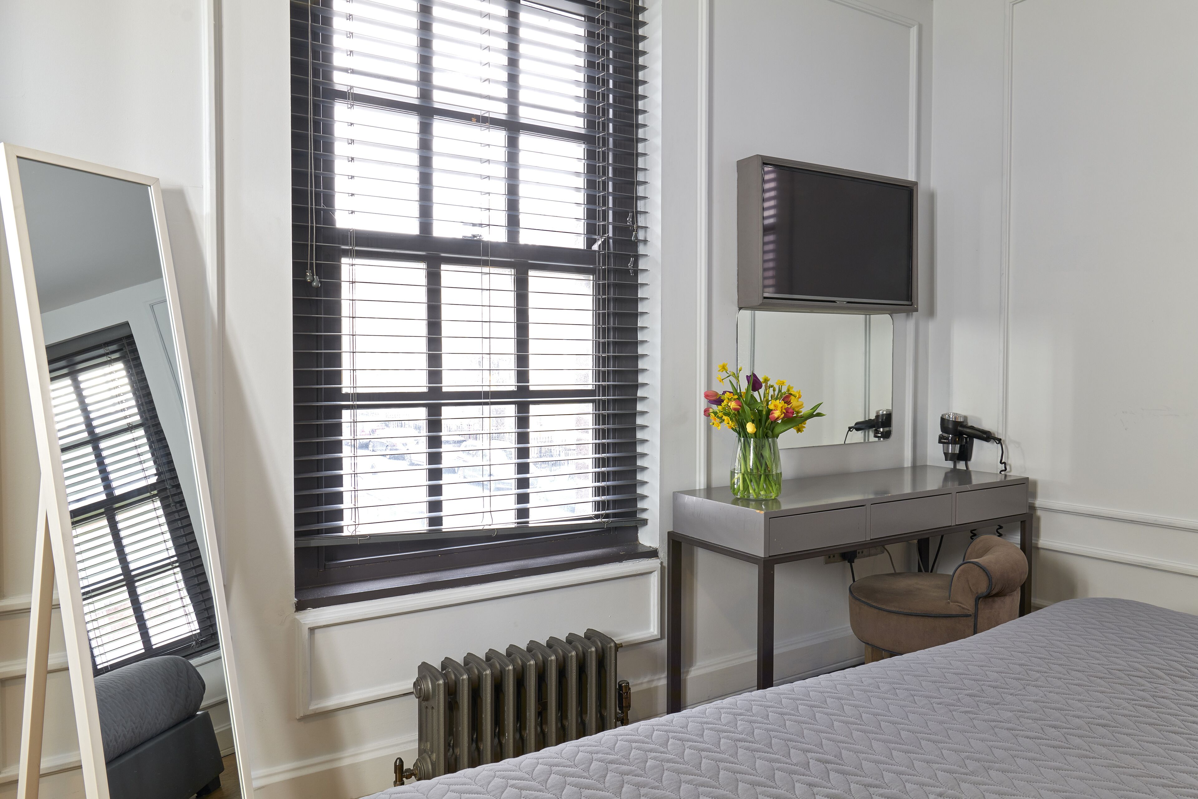 A bedroom with a bed, a dresser, a mirror, a TV, and a window with blinds.