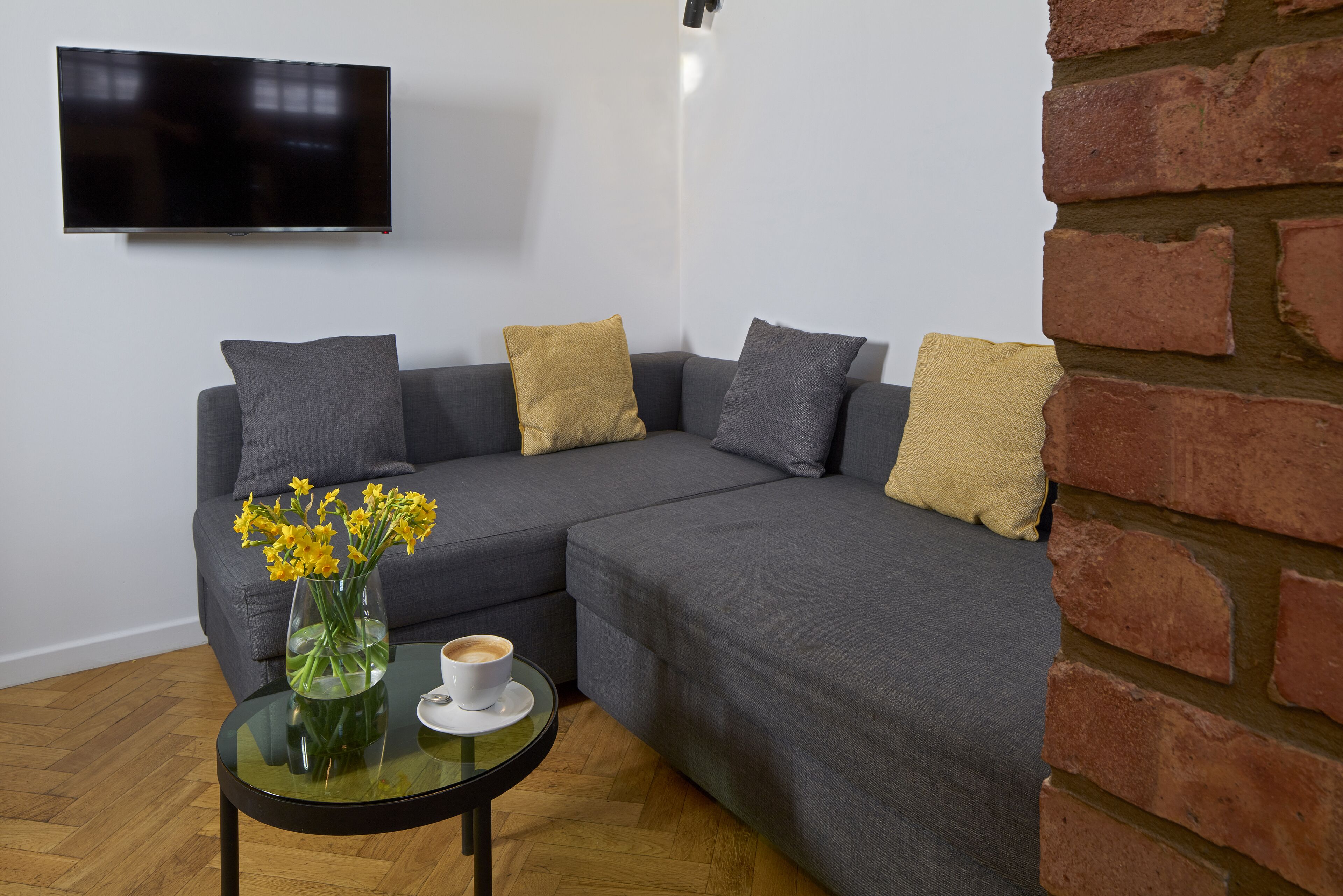 A modern living room with a grey sectional sofa, a glass coffee table with a vase of yellow flowers, and a flat-screen TV mounted on the wall.