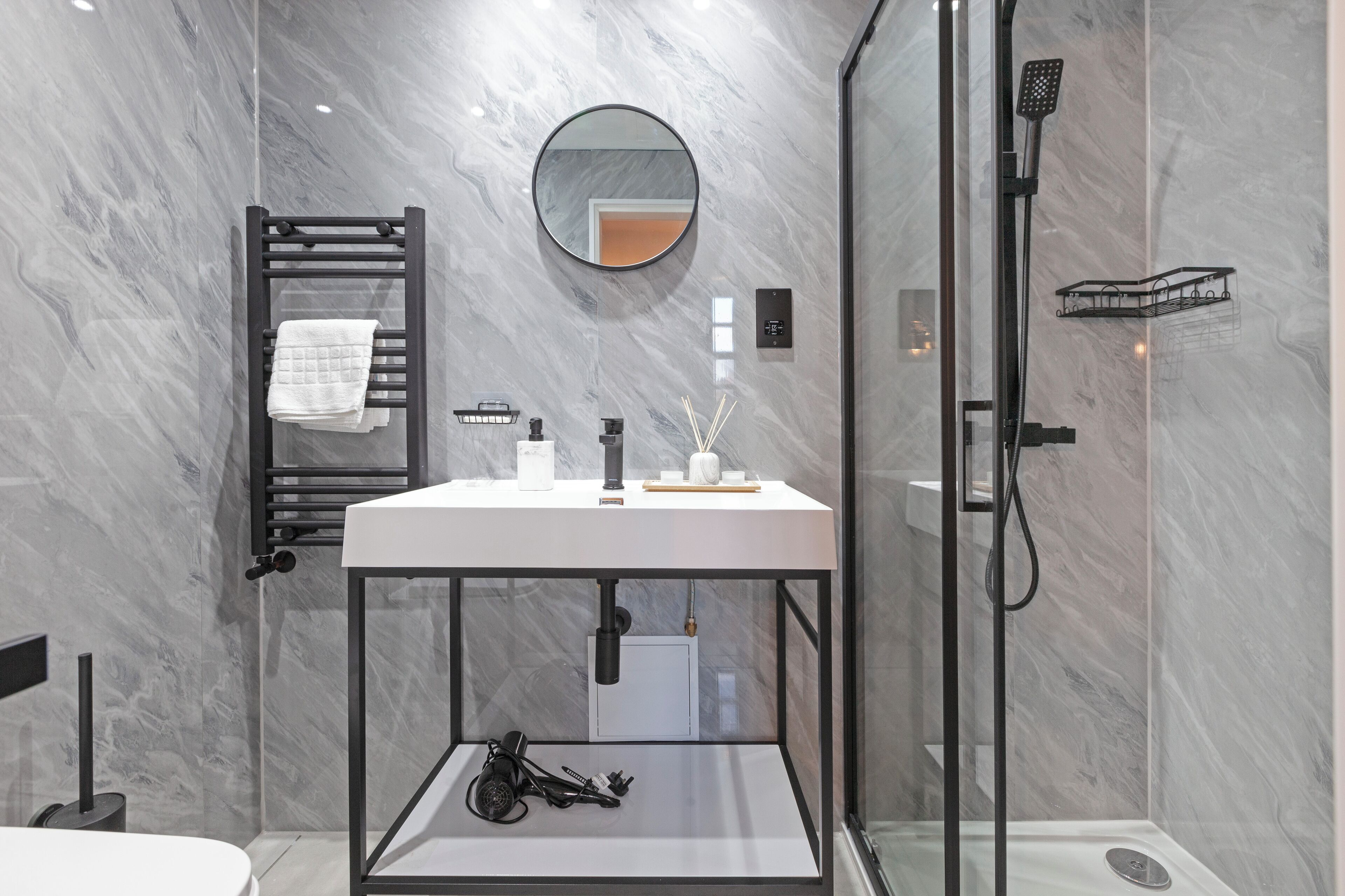 Une salle de bain moderne avec un mur en marbre, un miroir rond, un lavabo blanc et une douche séparée par une paroi de verre.